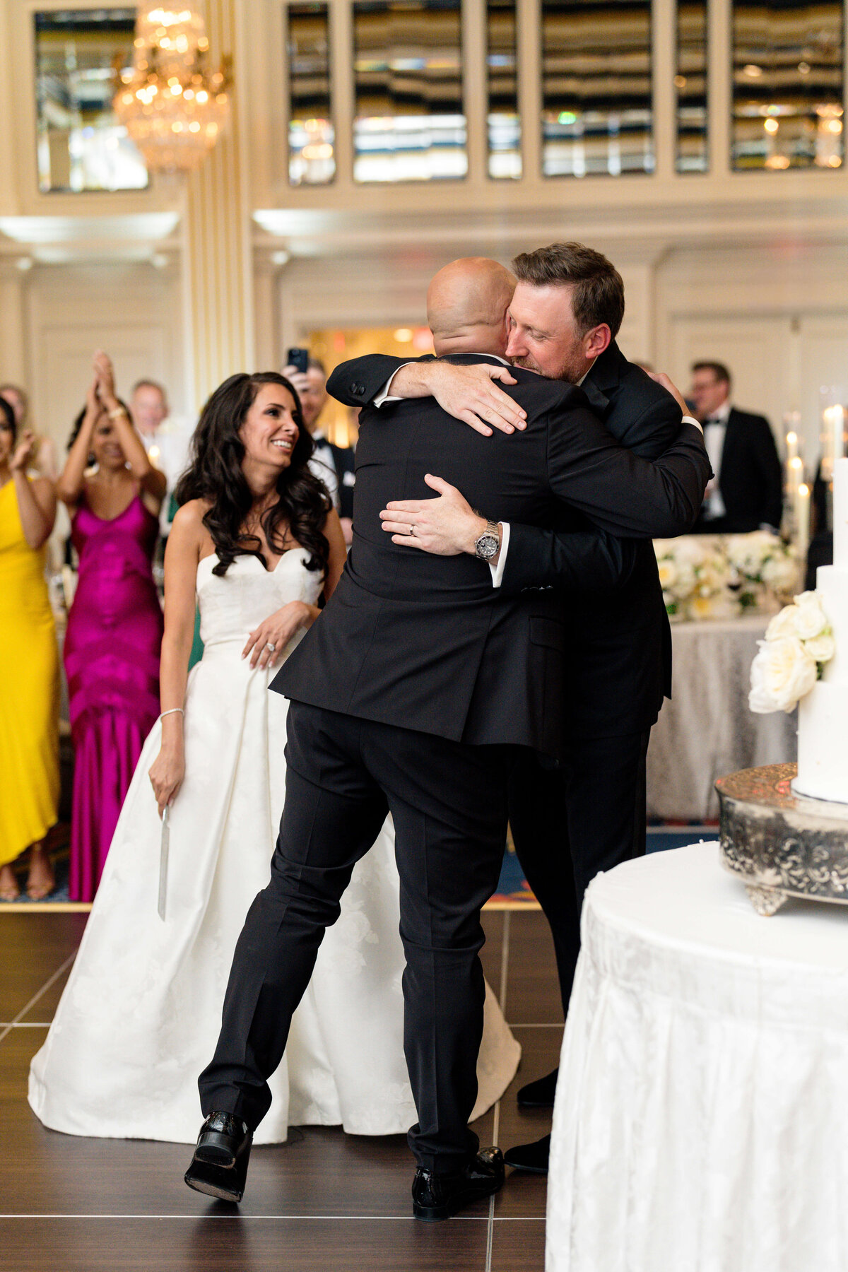 Two men in suits hug joyfully on the dance floor at a wedding reception. A woman in a white dress and another in a magenta dress cheer and clap in the background. The room is elegantly decorated with chandeliers and floral arrangements.