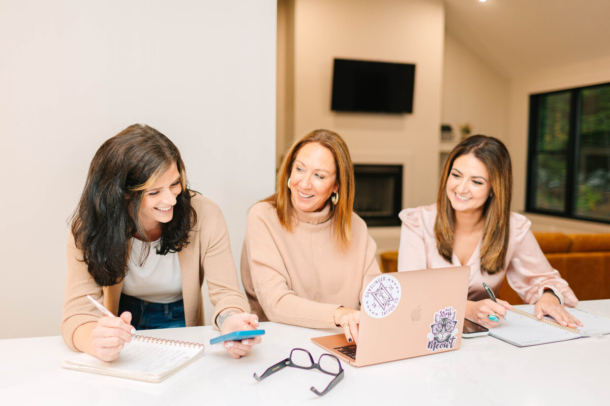 Three professional women sit at a white table, engaged in a collaborative discussion. One types on a rose gold laptop, another holds a smartphone, and the third takes notes. They are dressed in neutral and blush tones, with a modern living space featuring a fireplace and large windows in the background.