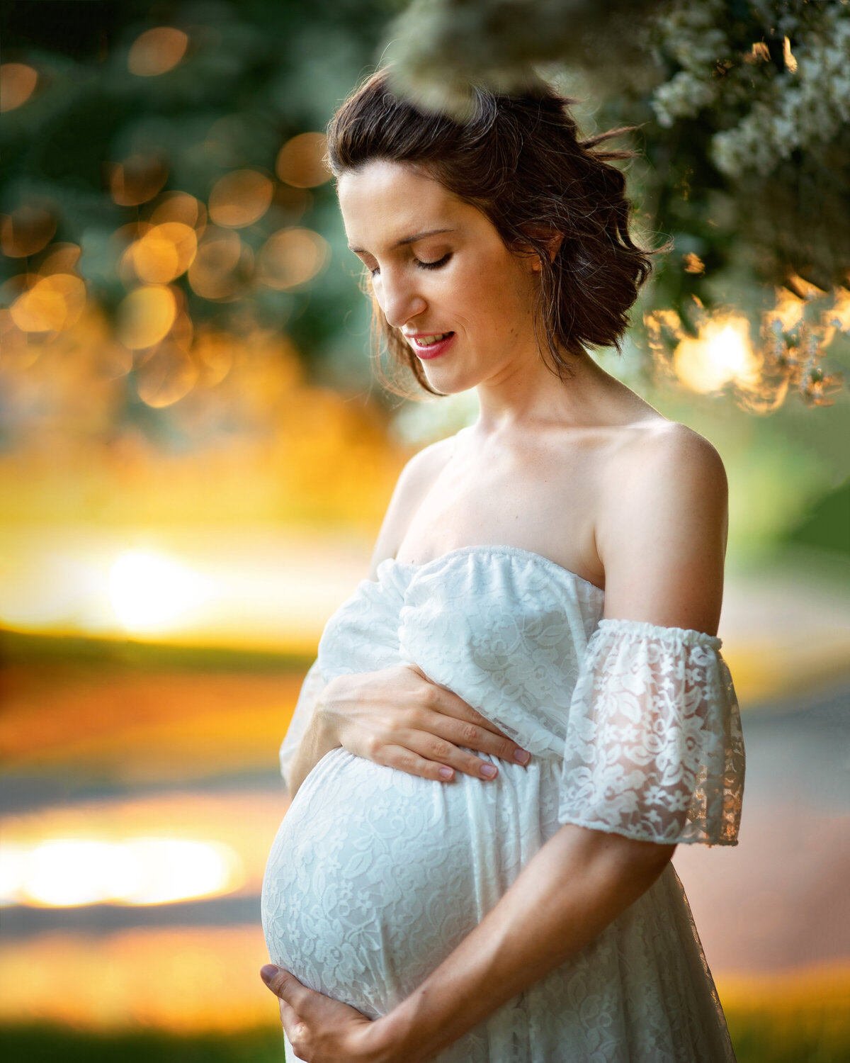 Woman with dark, short hair holding her pregnant belly.  She is smiling and standing under a crepe myrtle tree.  She is wearing a white dress and it is during golden hour.