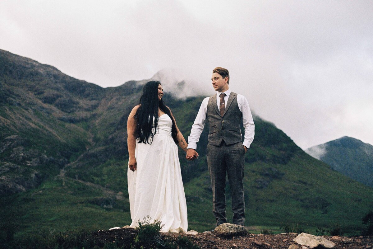 A bride and groom stand hand in hand in the Aberdeenshire landscape. The photography is grainy and captured on 35mm film. They wear wedding attire.