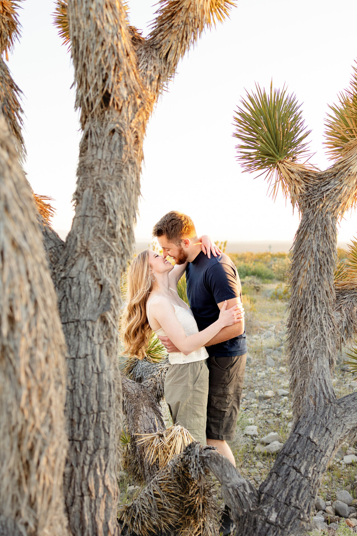 A couple shares a romantic moment framed between Joshua trees in the desert, wrapped in each other’s arms as the sun sets in the distance.