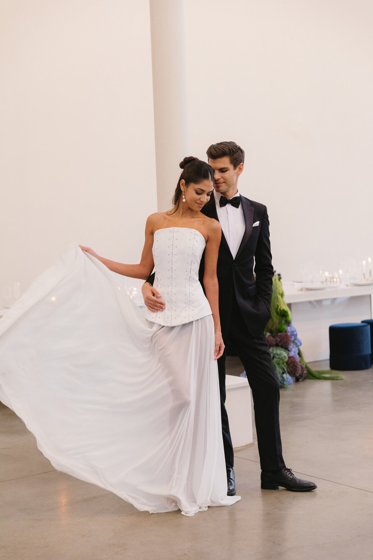 Bride creating movement with her flowing dress while groom stands behind her in an elegant contemporary wedding editorial.