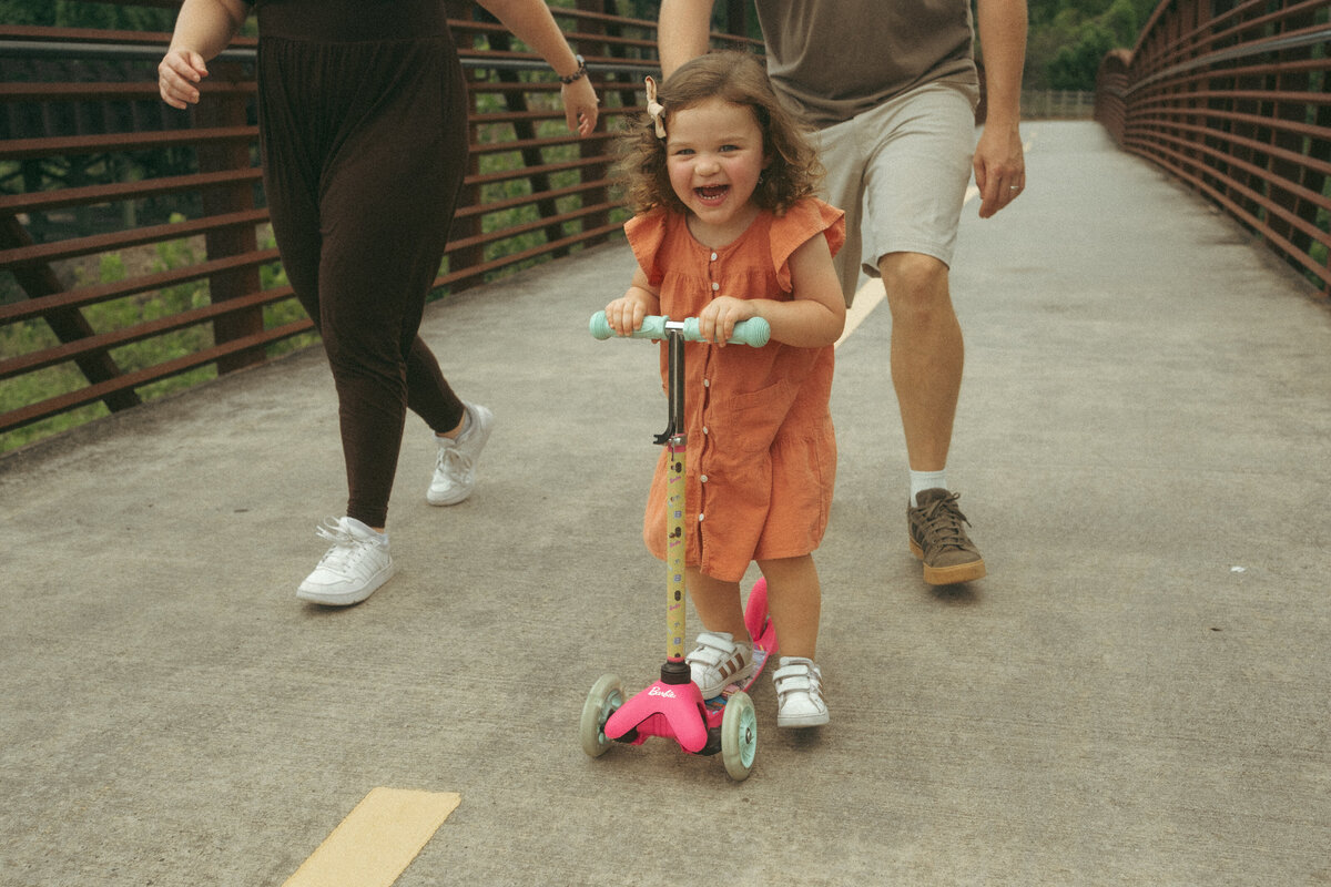 A happy toddler girl on a bright pink scooter riding in Noonday Creek Trail while her parents walk behind her. 