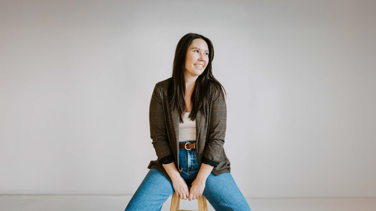 Headshot of Colorado wedding and portrait photographer Avenir Photo Co. sitting on a stool in a minimalist studio, smiling naturally in soft light.
