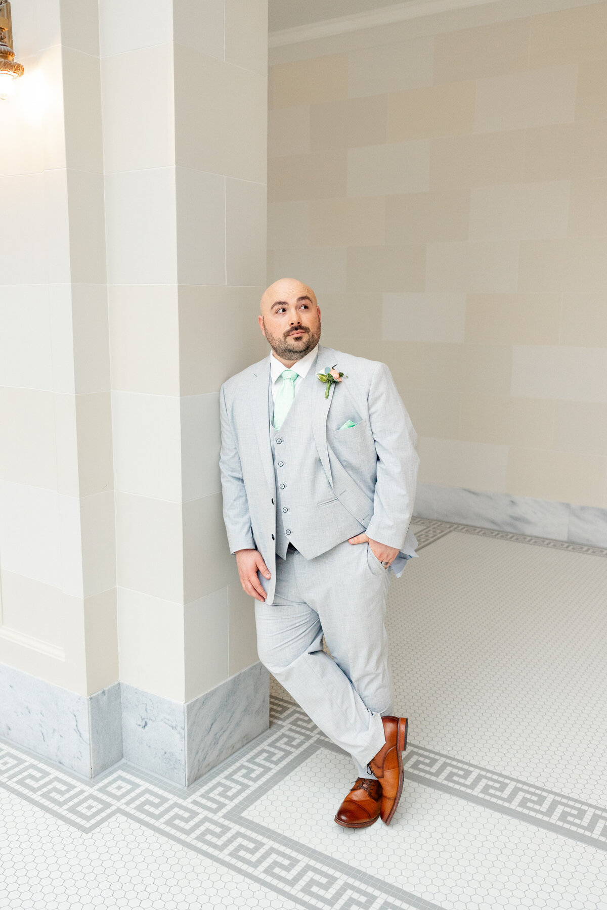 The groom, dressed in a three-piece gray suit, leans against a marble column in the Utah State Capitol, looking off into the distance.