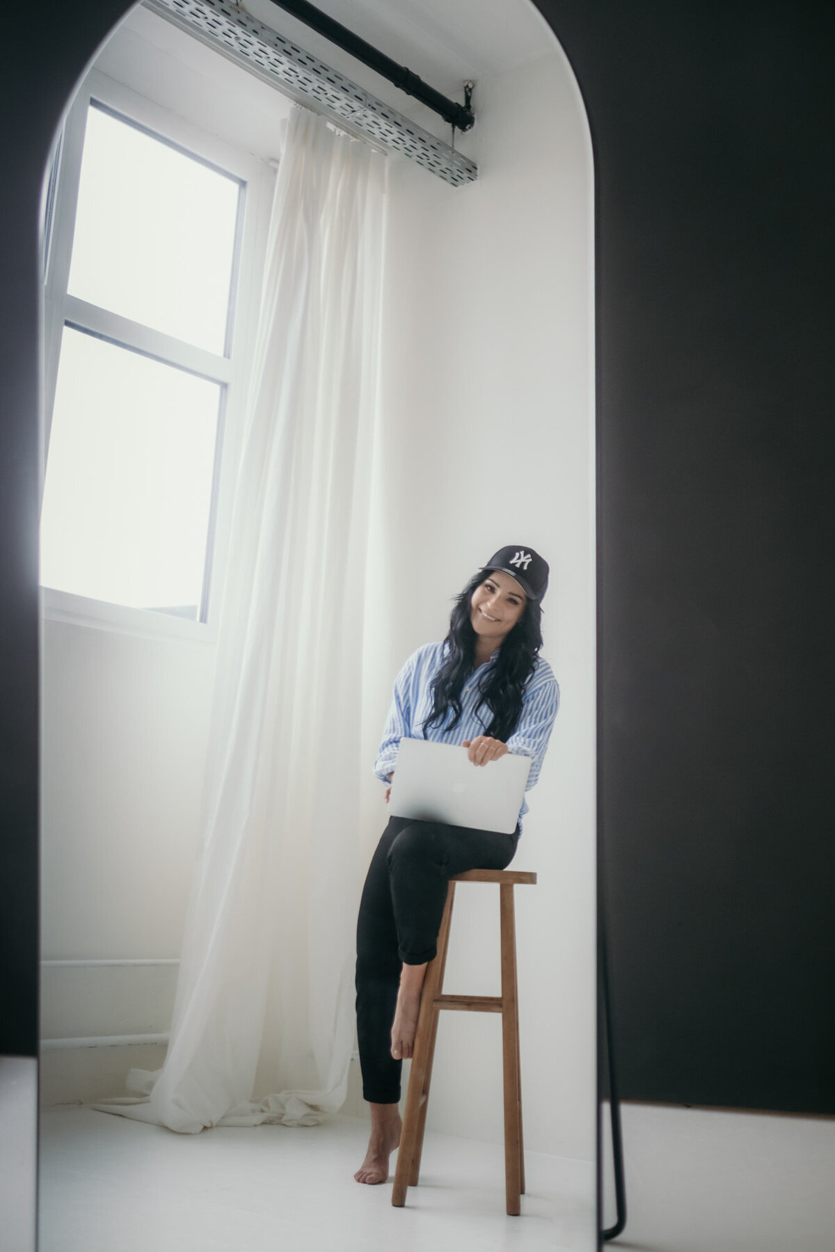 Woman sits on a stool besides a window working on her laptop.