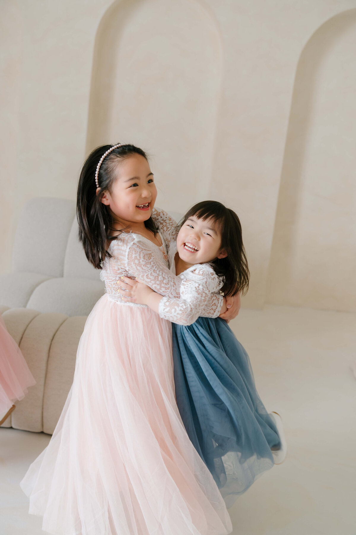 Two sisters dance and hug in matching lace dresses during a lighthearted family studio moment.