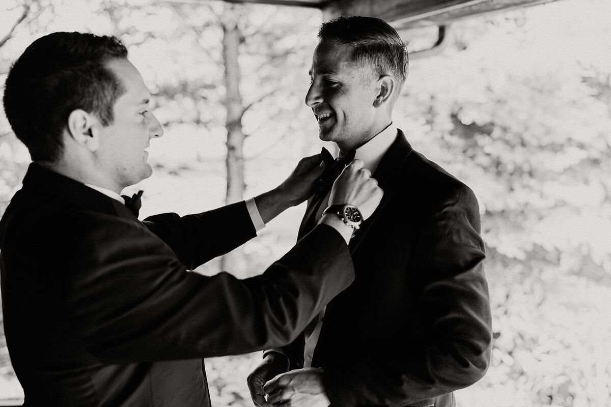 black and white image of a groom having his tie adjusted by a groomsman on the porch of the preparation cottage on the grounds of Willowbrook wedding venue