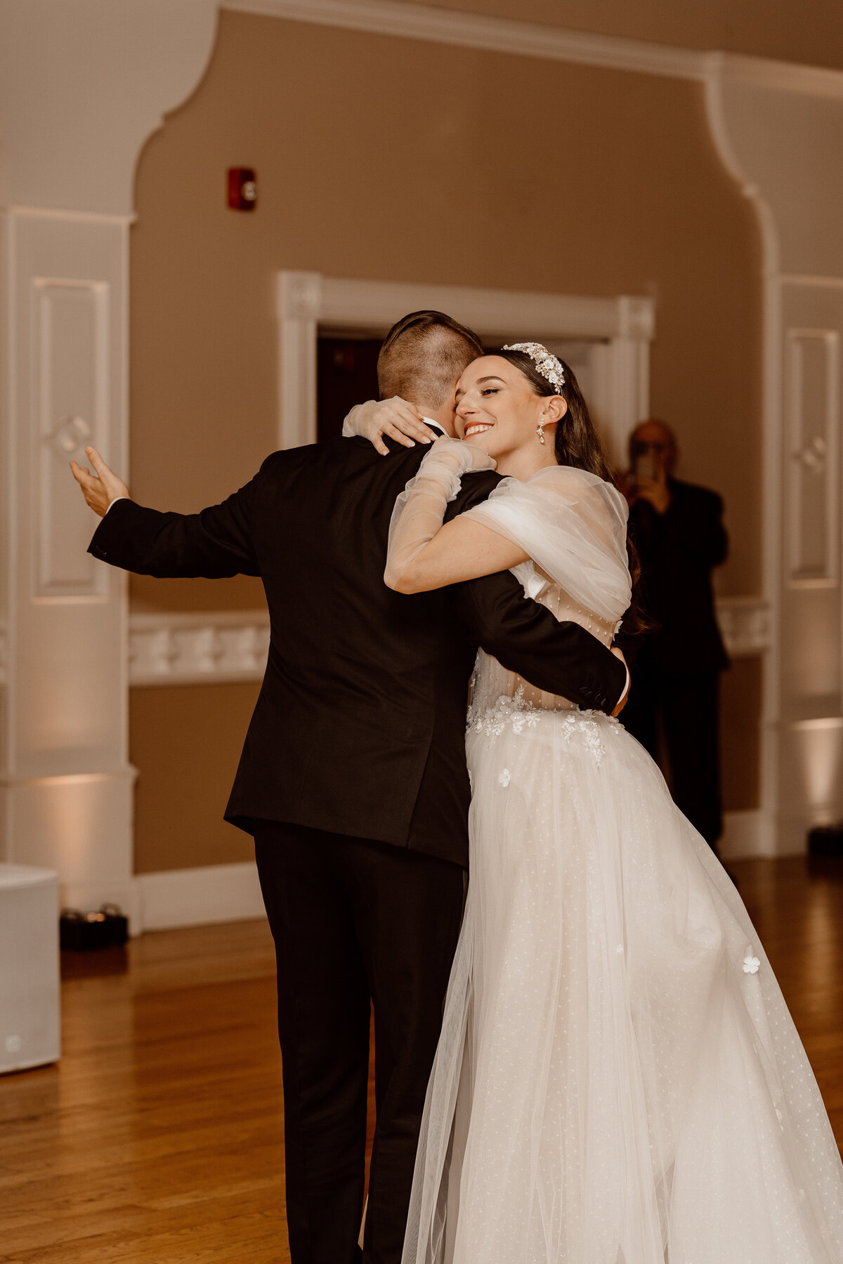 Bride and groom share a heartfelt embrace during their first dance at the wedding reception. The bride, in a delicate tulle gown with floral details and a sparkling headpiece, smiles radiantly. The groom, in a classic black suit, holds her close in a beautifully decorated venue with soft lighting, capturing an intimate and joyous moment.