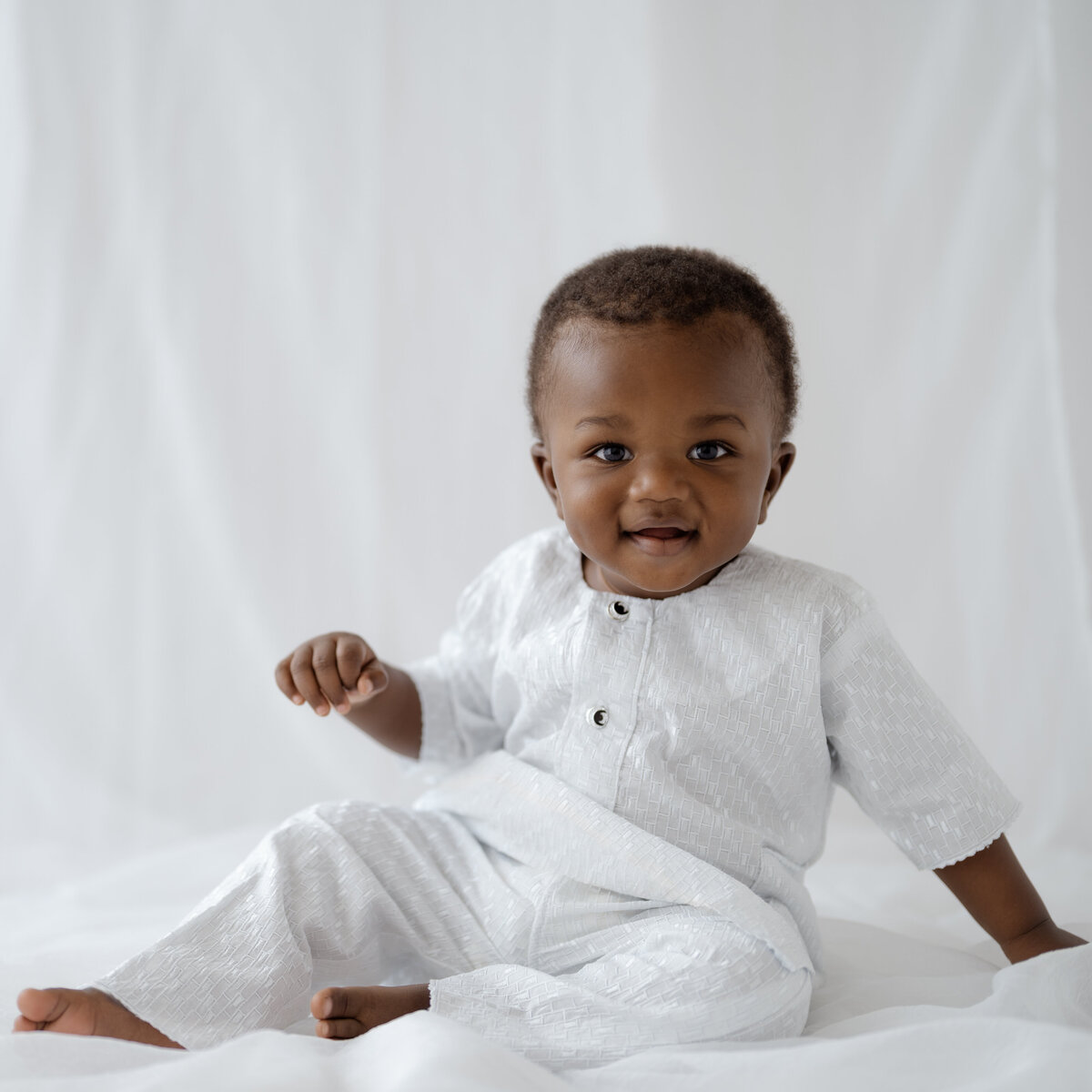 Adorable one-year-old boy posing for his milestone photoshoot in a Fort Worth studio. Dressed in a white traditional outfit, he smiles warmly while sitting against a soft white backdrop, capturing the joy and innocence of this special moment.