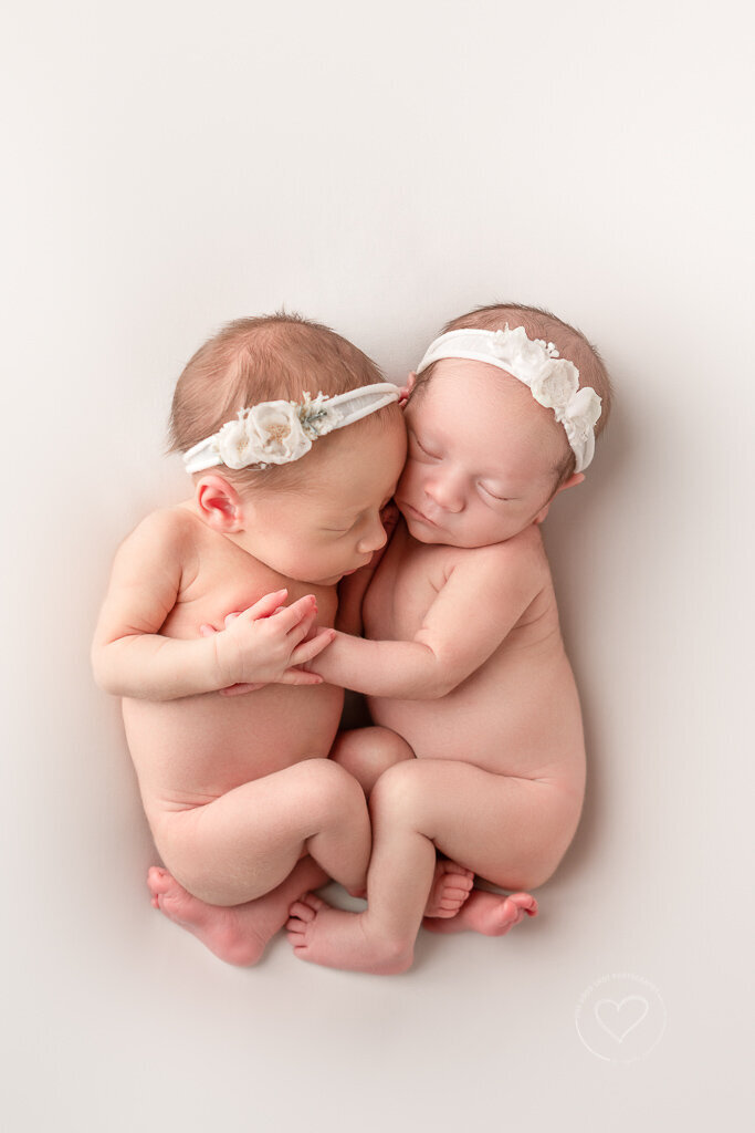 Twin baby girls curled together on all white backdrop with soft  lighting at One Good Shot Photography's Fresno CA studio