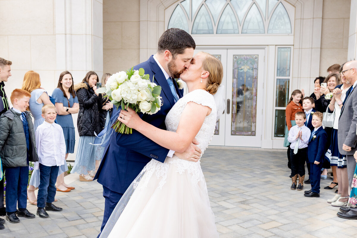 Close-up of bride and groom kissing as the groom holds the bride in his arms, surrounded by smiling guests and children outside the Taylorsville Utah LDS Temple.
