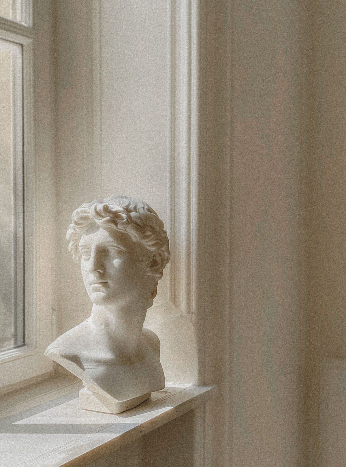 Bust sculpture placed on a deep windowsill, with soft light streaming into the white-painted room, creating a serene and sophisticated atmosphere.