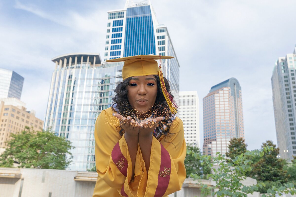 SENIOR IN CAP AND GOWN STANDING IN FRONT OF UPTOWN CHARLOTTE SKYLINE