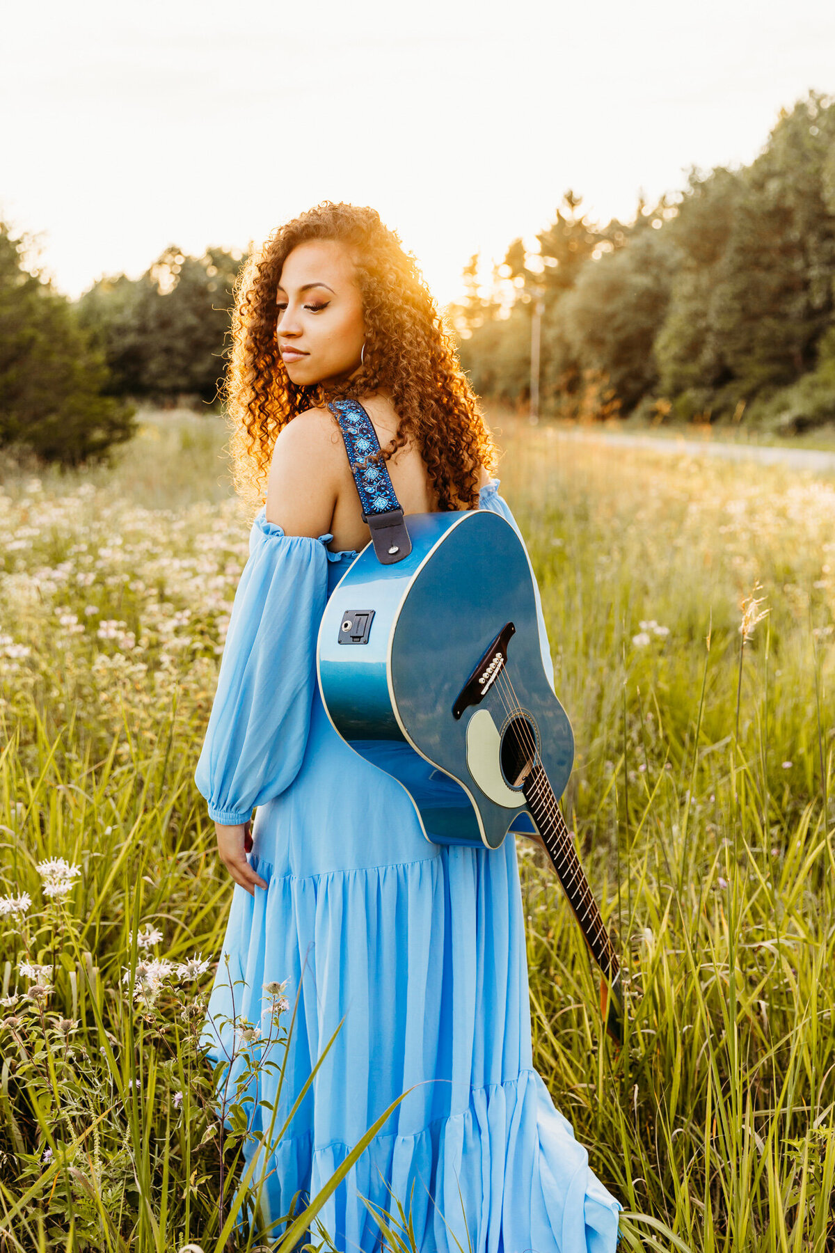 girl in blue dress with her guitar strapped to her back looking down at her shoulder