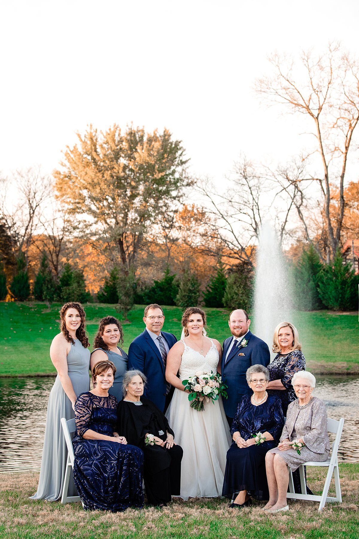 Bride in a sleeveless v-neck wedding dress and veil standing with the groom dressed in a navy blue suit with their family. The bridesmaids are dressed in a dusty gray blue. The mother of the bride and mother of the groom are dressed in midnight blue floor length dresses. The grandmothers of the of the bride and groom are dressed in navy blue and silver.