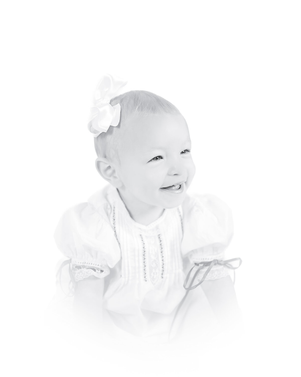 Black and white heirloom portrait of a smiling baby dressed in a classic white outfit with lace details. This timeless baby photography captures the innocence and joy of childhood, created by a Birmingham newborn photographer specializing in elegant, light-filled portraits.