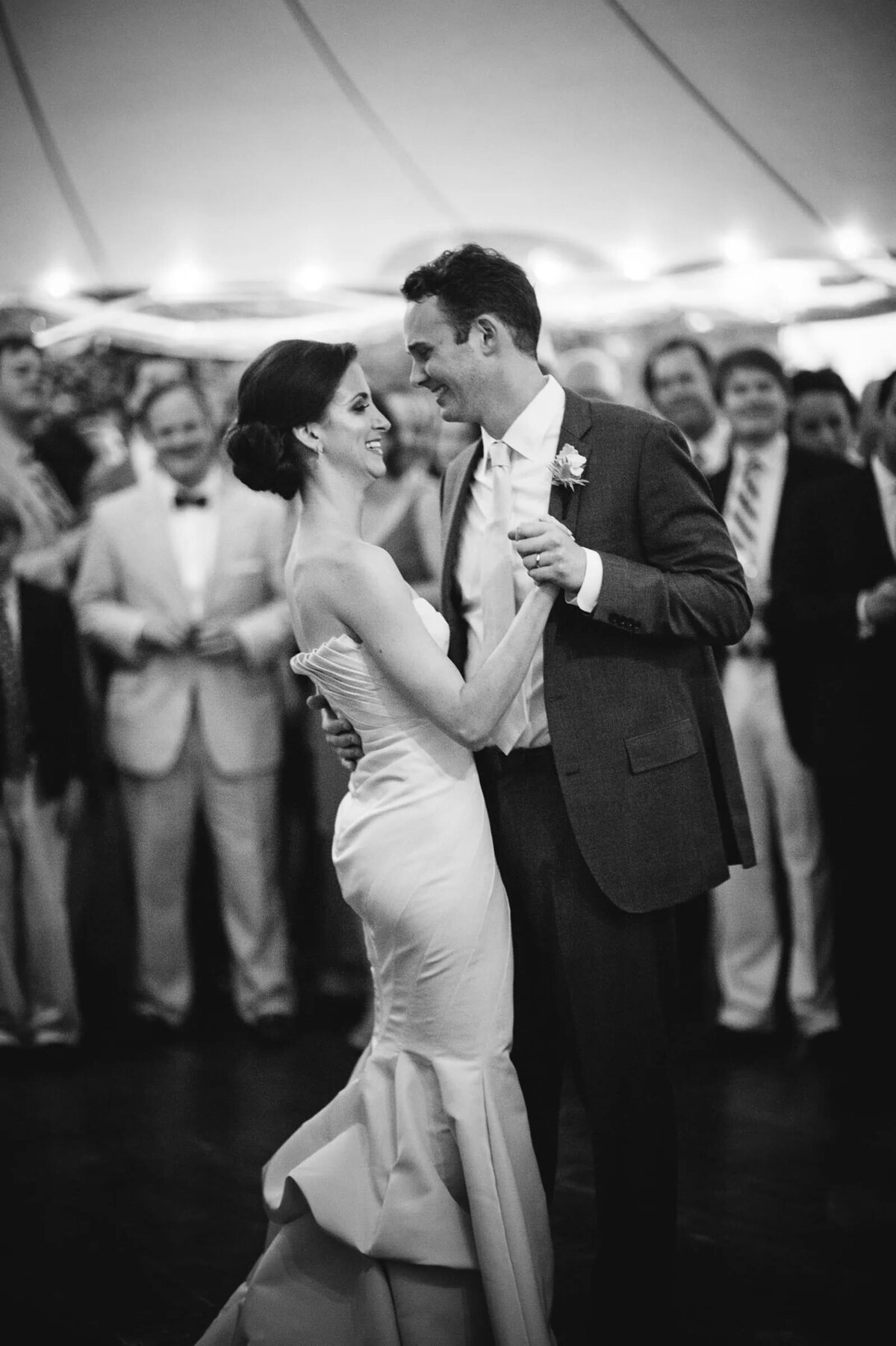 A couple dances intimately on their wedding day, the bride laughing as the groom holds her close, with guests surrounding them in a candid moment.