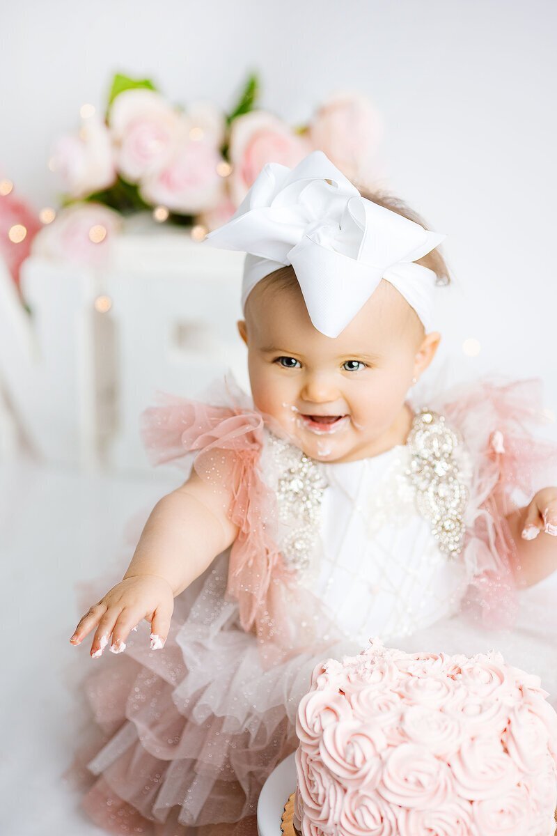Baby wearing a big bow and pink ruffled dress smiling and holding cake, taken by photographers new jersey Ally Kimpton Photography