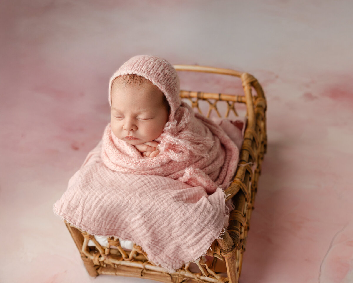 Newborn baby girl wrapped in soft pink tones, peacefully resting in a woven basket with a matching bonnet. Captured during her Inland Empire newborn session by Kaitlyn Dawn Photography.