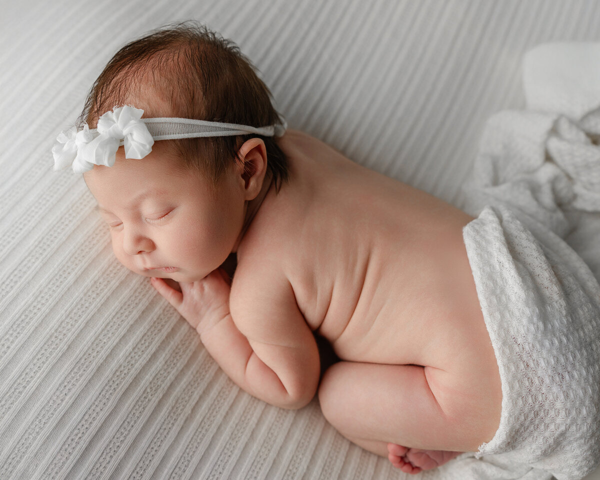 Sweet newborn baby girl with a white floral headband, peacefully sleeping on her tummy, wrapped in a soft textured blanket. Captured by Inland Empire newborn photographer Kaitlyn Dawn Photography.
