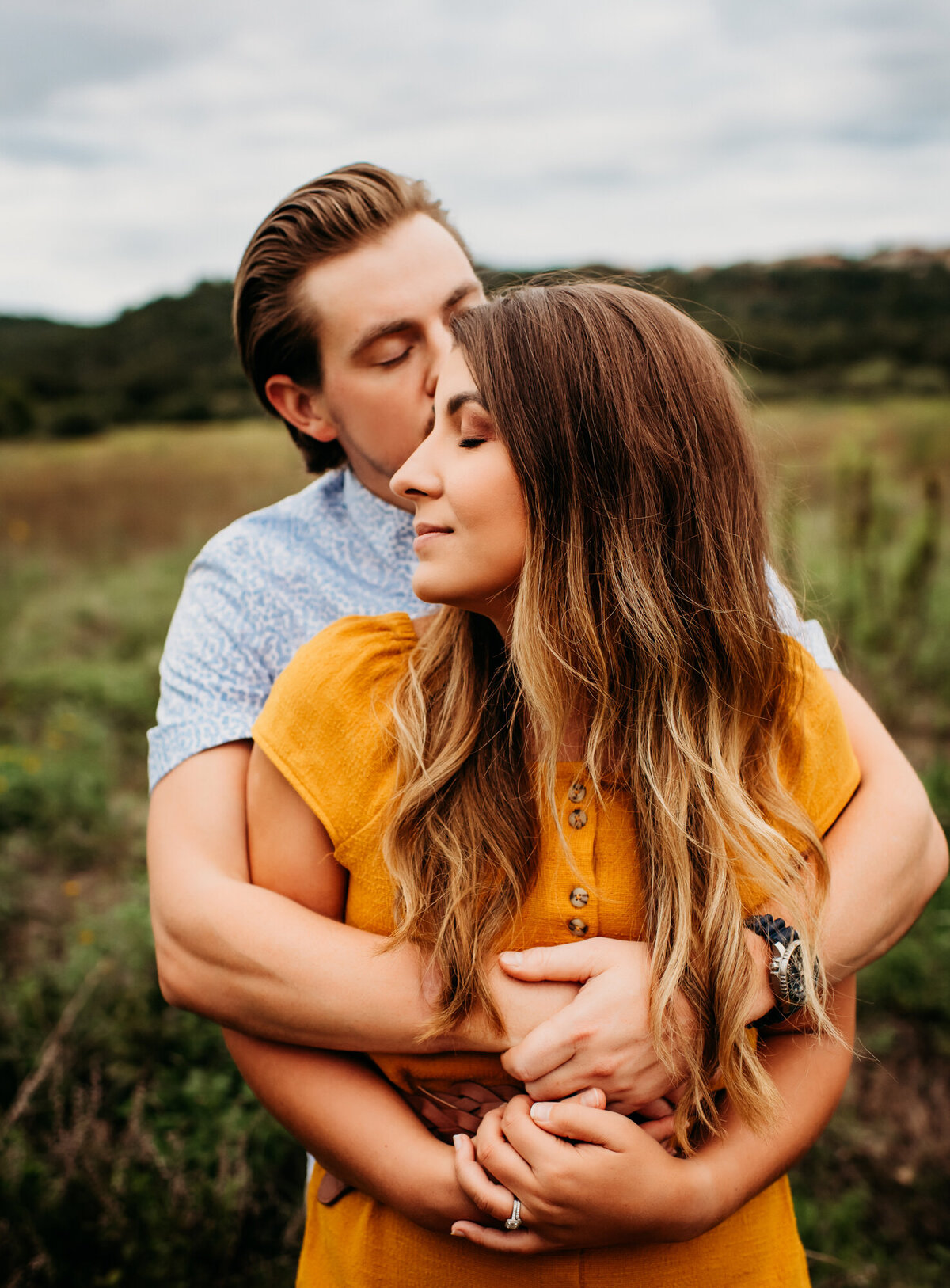 Couples Photography, Man in a blue t shirt wraps his arms around a woman in a gold dress and kisses her on the side of her face.