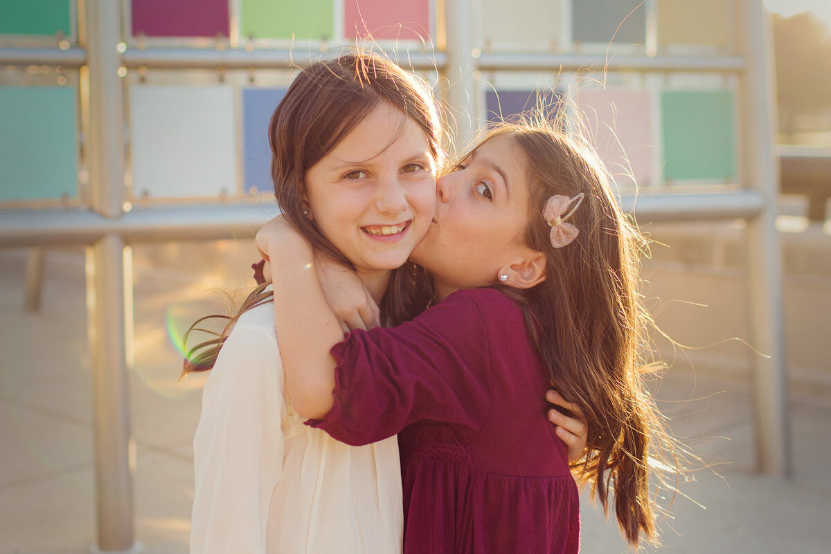 Younger sister hugs and gives older sisters a big kiss on her cheek. Colorful tiles are behind them.