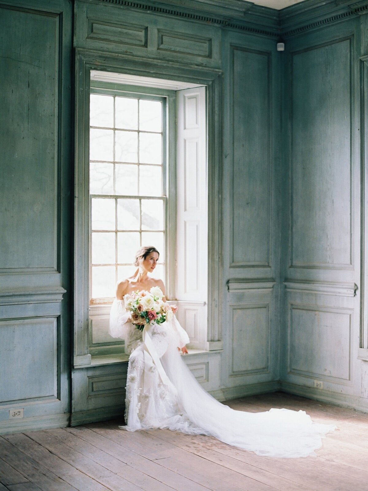 Newport Wedding Photographer | A bride in an elegant white gown holds a bouquet while seated by a large, bright window in a softly lit, vintage-style room.