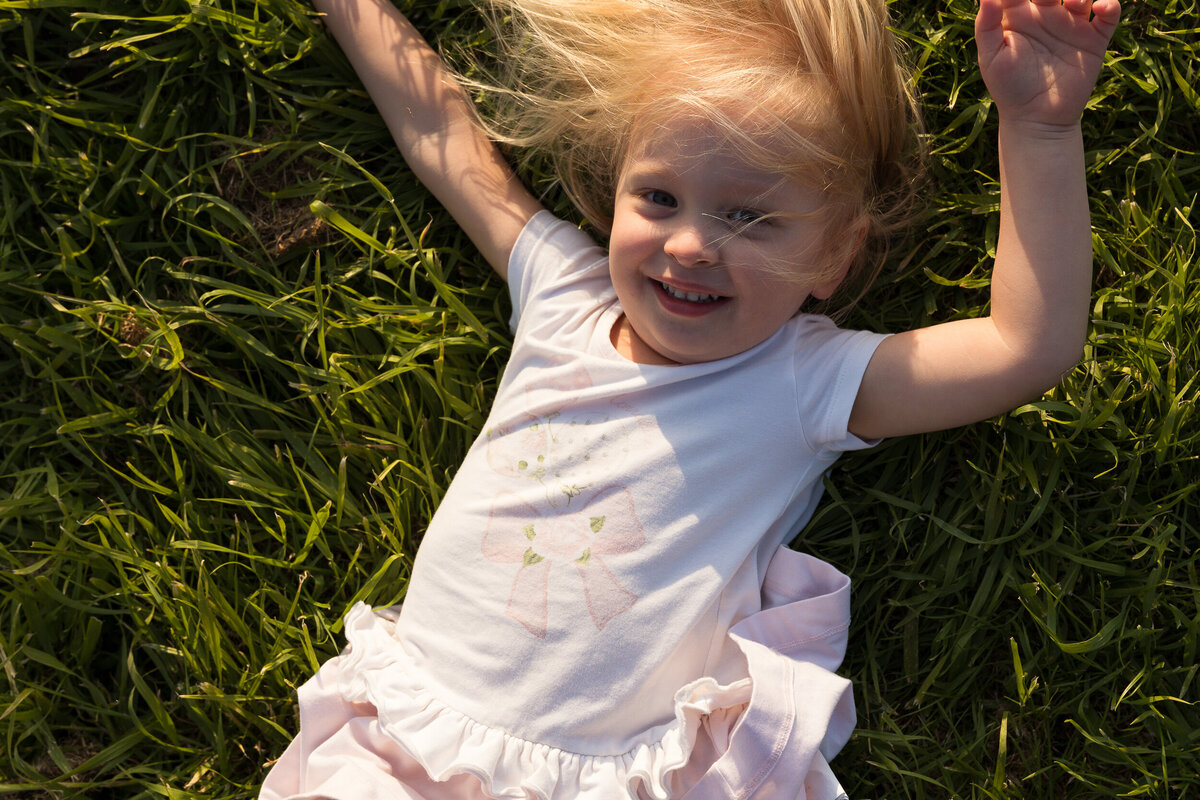 A young girl with blonde hair, wearing a pink skirt and white top, lying and smiling in the grass.