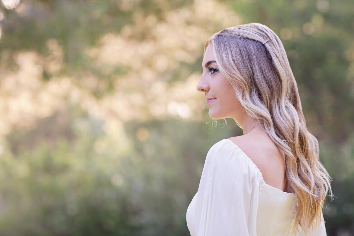 A woman with long, wavy blonde hair stands outdoors in a serene, blurred background of greenery. She is wearing a white, off-the-shoulder top and is looking to her right.