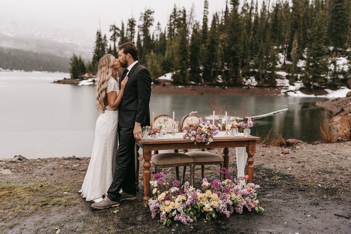 A couple leans together on a decorated table in front of a frozen lake. The table is decorated in gorgeous florals.