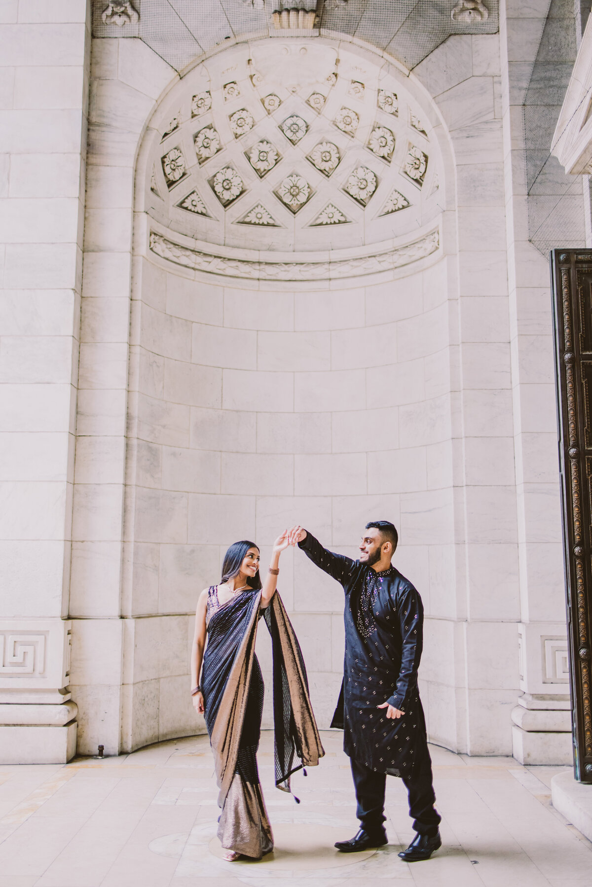an engaged couple in traditional indian black clothes, sari and kurta, twirling outside the alcove at the new york public library