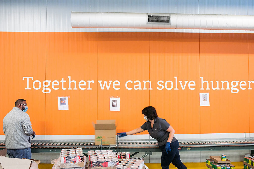 Two people work on a conveyor belt with text on the wall that says  "Together we can solve hunger."