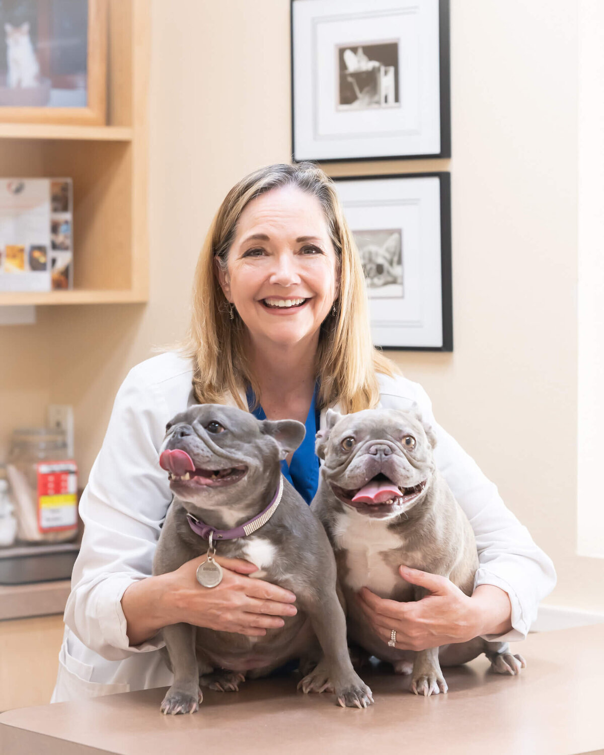 Vet clinic owner with her two frenchies, smiling, with her arms around them in an exam office