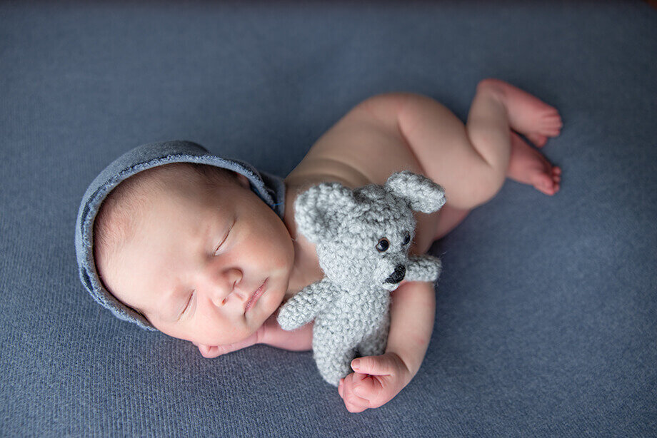 newborn boy posed side lying on top of denim blue fabric.  He is holding a grey teddy bear and wearing a matching blue beanie