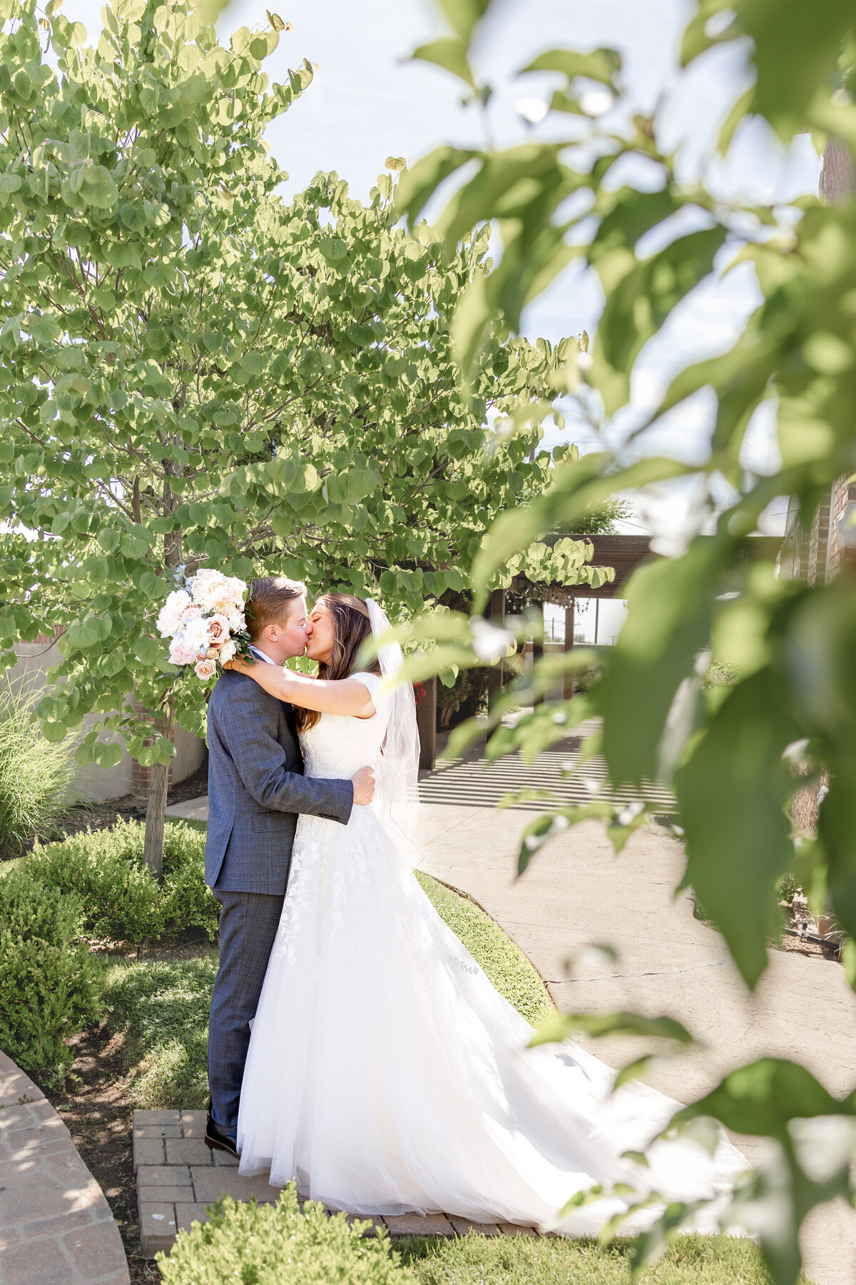 A newlywed couple shares a passionate kiss outdoors at the Talia Event Center, framed by lush greenery.