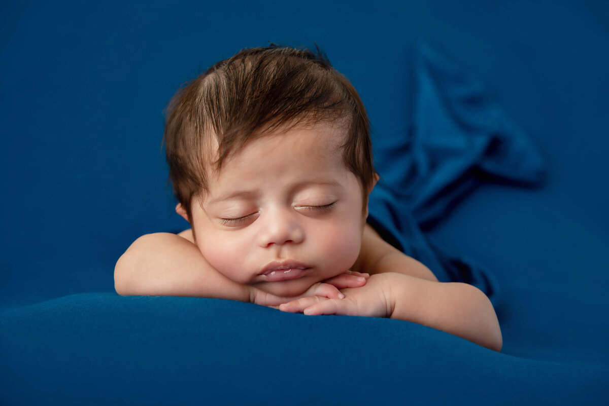 newborn baby boy laying on a blue background with his head on his hands for his newborn photos