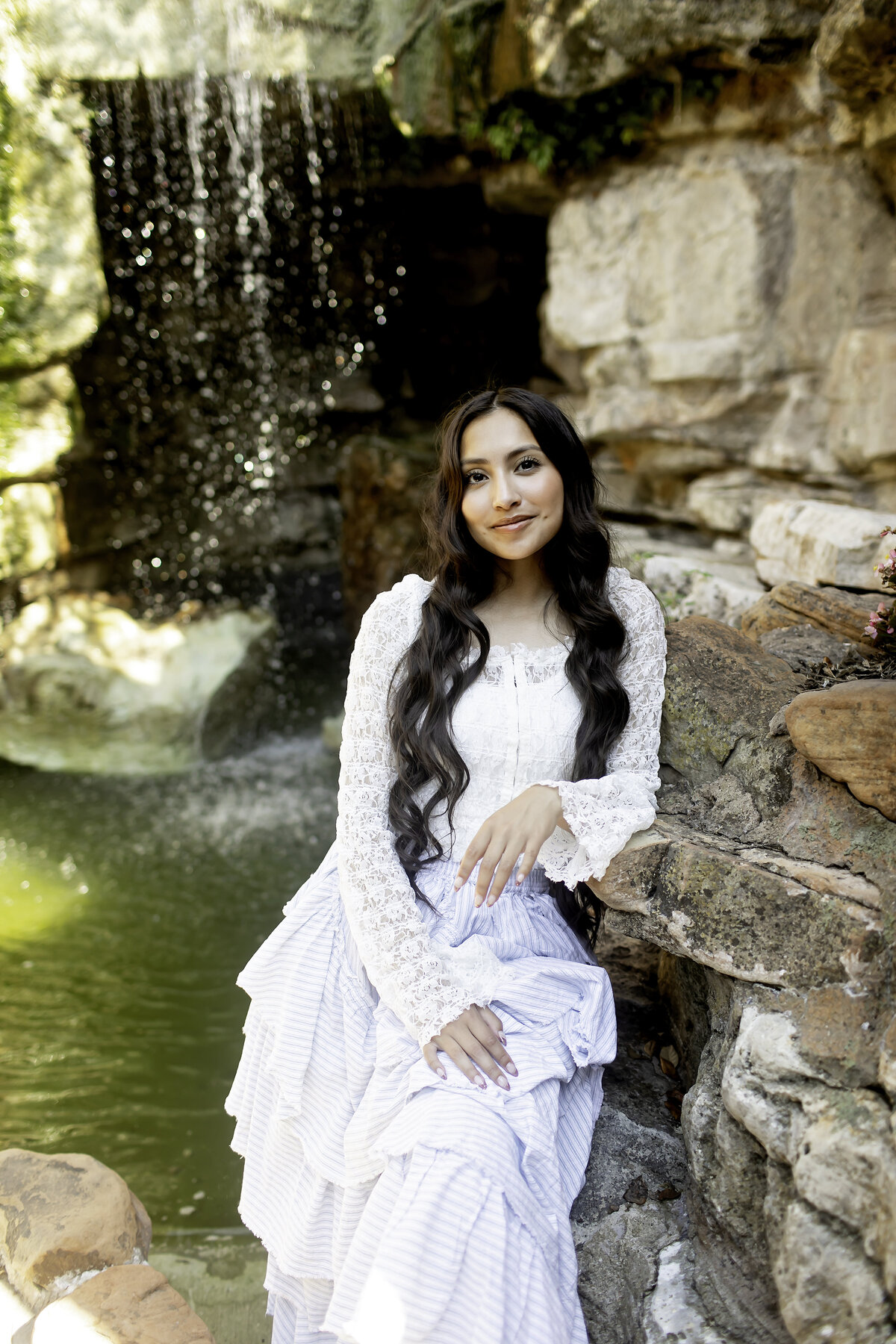 A high school senior standing near a beautiful stone fountain at Chandor Gardens in Weatherford, TX, with the garden's serene ambiance adding to the elegance of her photoshoot