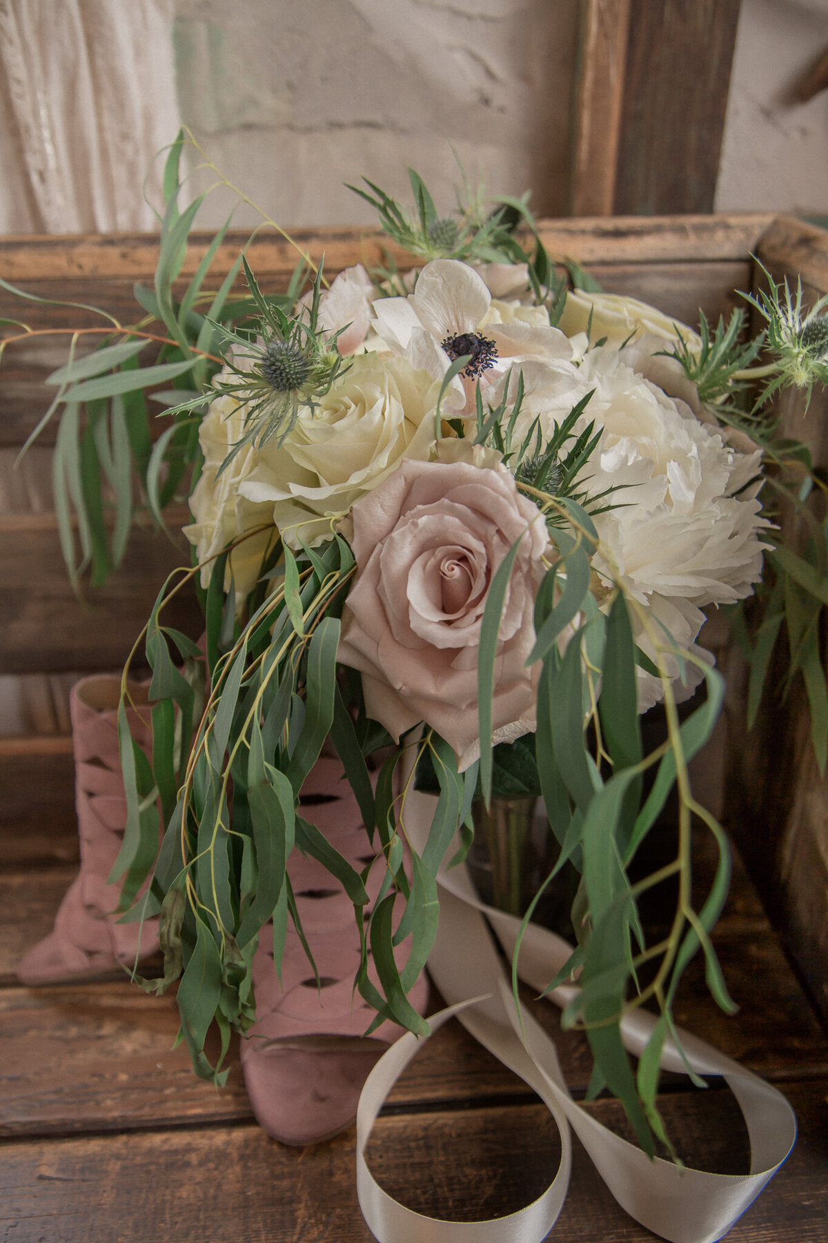 The bridal bouquet of roses, anemones and eucalyptus rest beside the brides pink heeled sandals