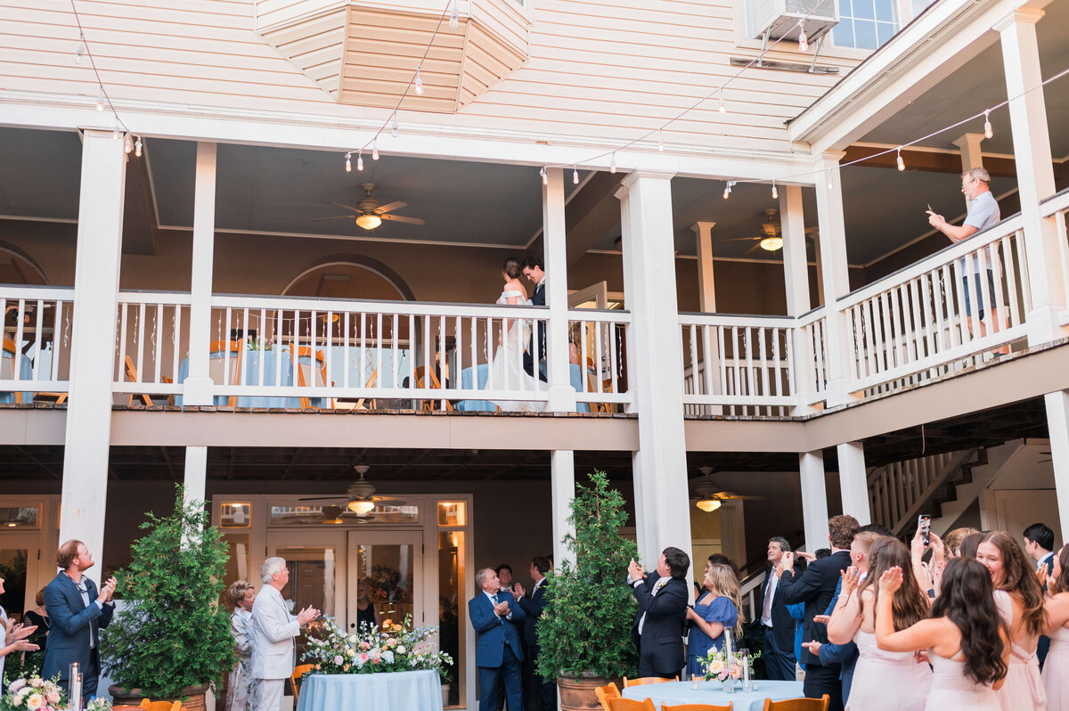 A bride and groom walk along an upper balcony at an outdoor wedding venue as guests below look up, cheer, and take photos. The scene is festive, with tables, flowers, and string lights.
