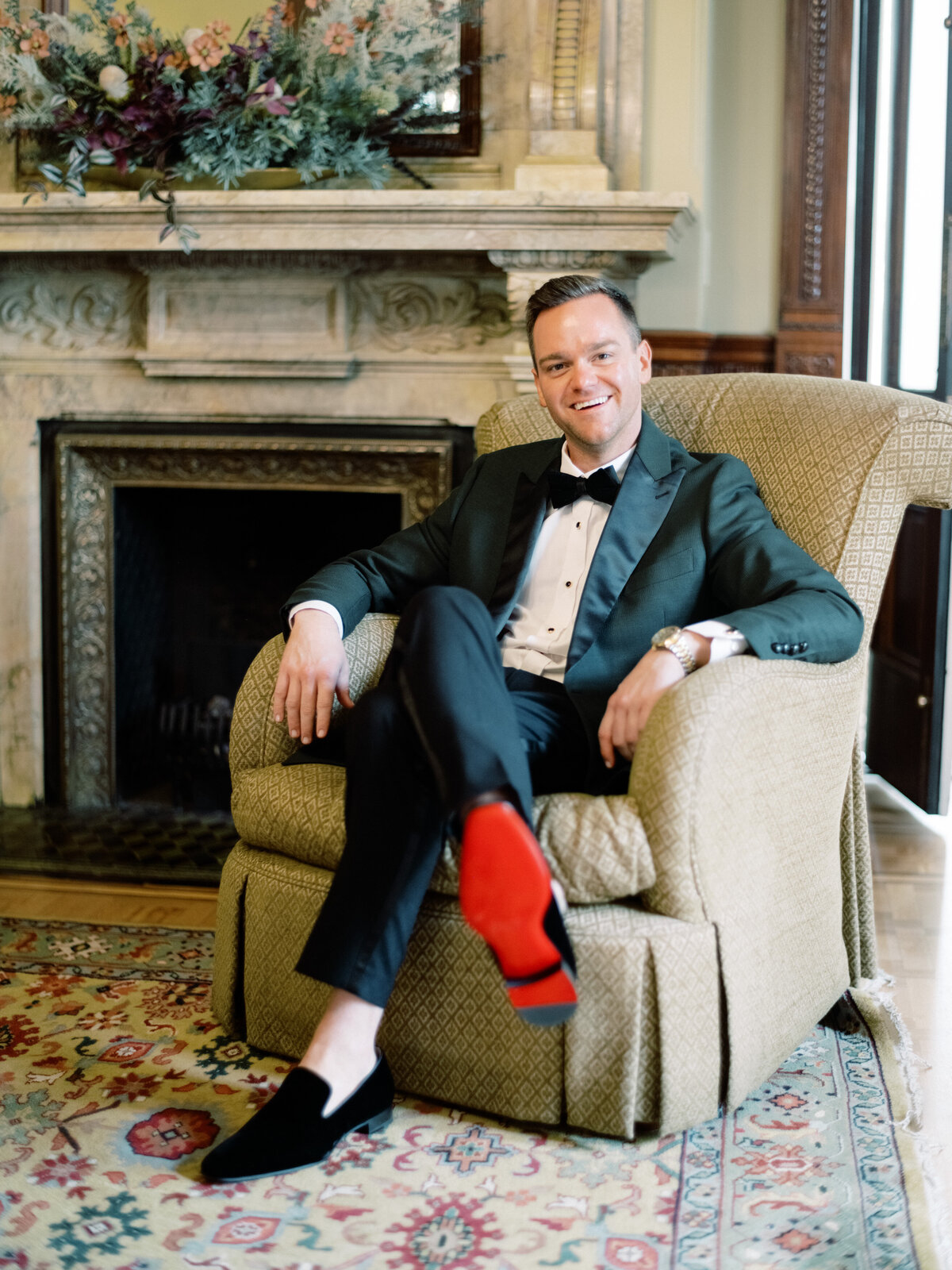 A stylishly dressed man sits comfortably in an elegant armchair, showcasing his black tuxedo and distinctive red-soled shoes in a beautifully decorated room.