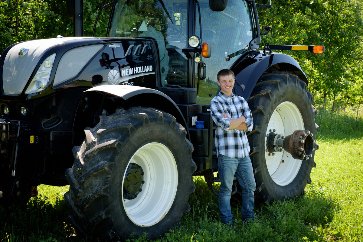 Luxemburg Casco high school senior boy wearing a black and white plaid button down shirt and jeans by a black New Holland tractor in farm field at his home near Green Bay, Wisconsin.