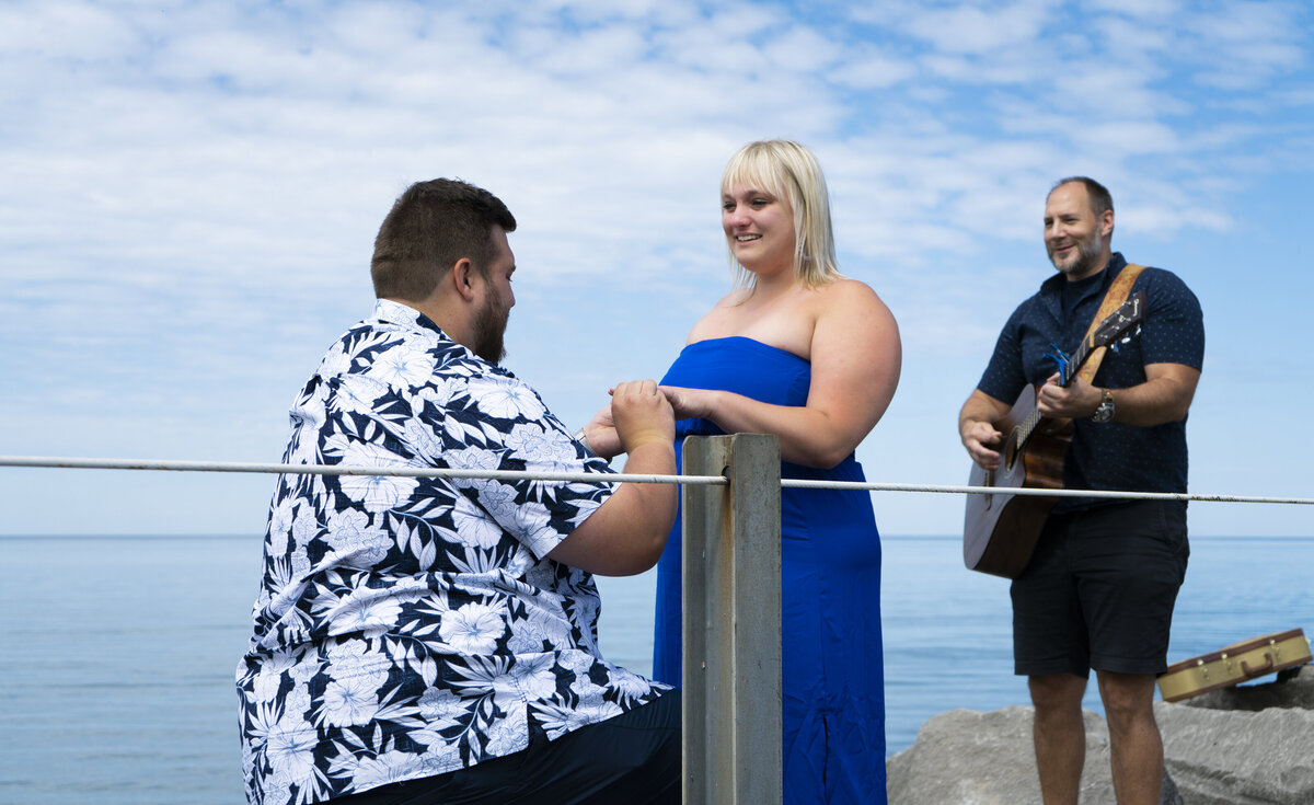 Photo of a man proposing to his girlfriend near a beach with a guitarist
