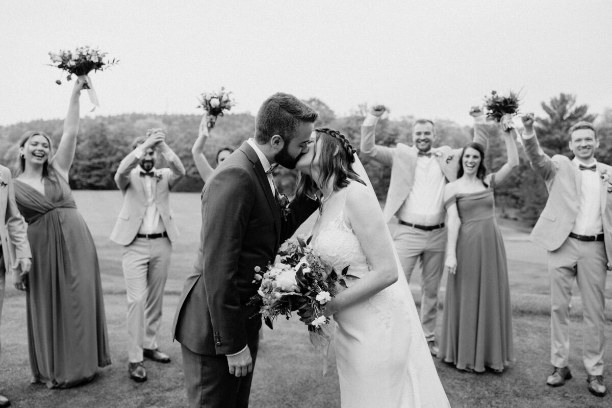 A bride and groom kiss while the bridesmaids and groomsmen cheer at the back