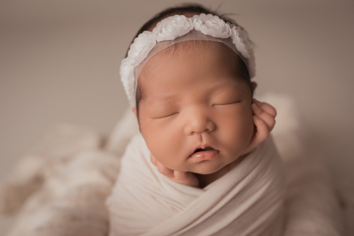 newborn portrait session at atlanta ga maternity and newborn photography studio casey mcminn photography with baby girl in white flower headband, cream swaddle and hands on cheeks