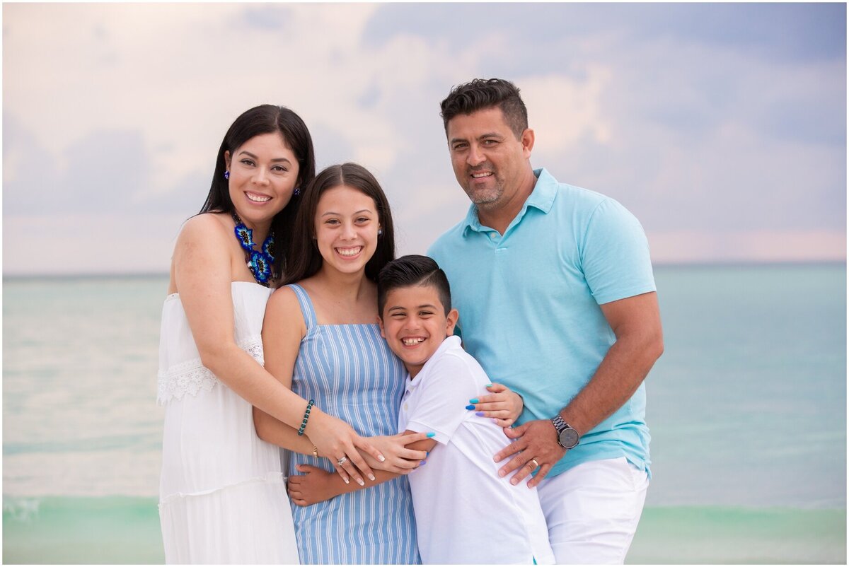 Family wearing neutral colors hugging  and smiling for the camera on the beach