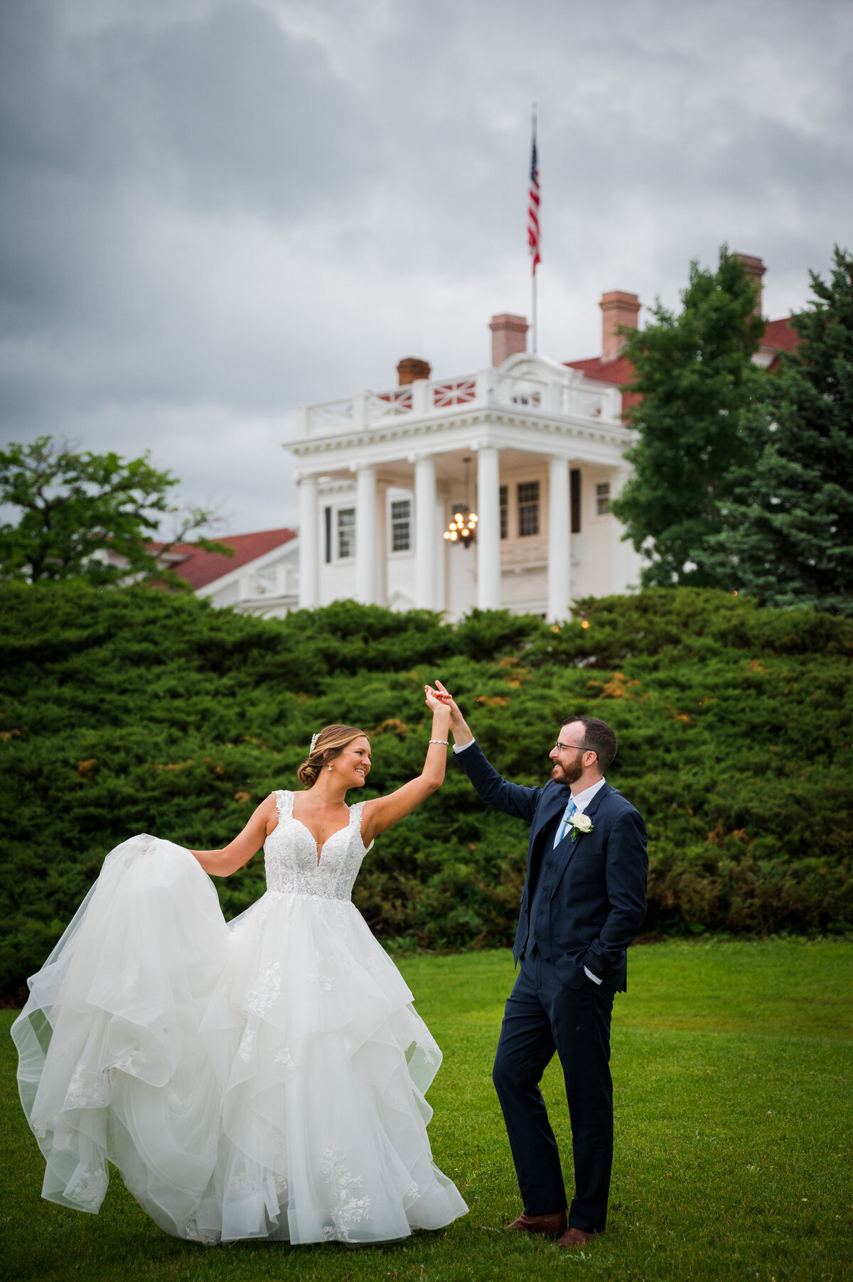A groom spins his bride as she holds her dress with The Manor House in the background.