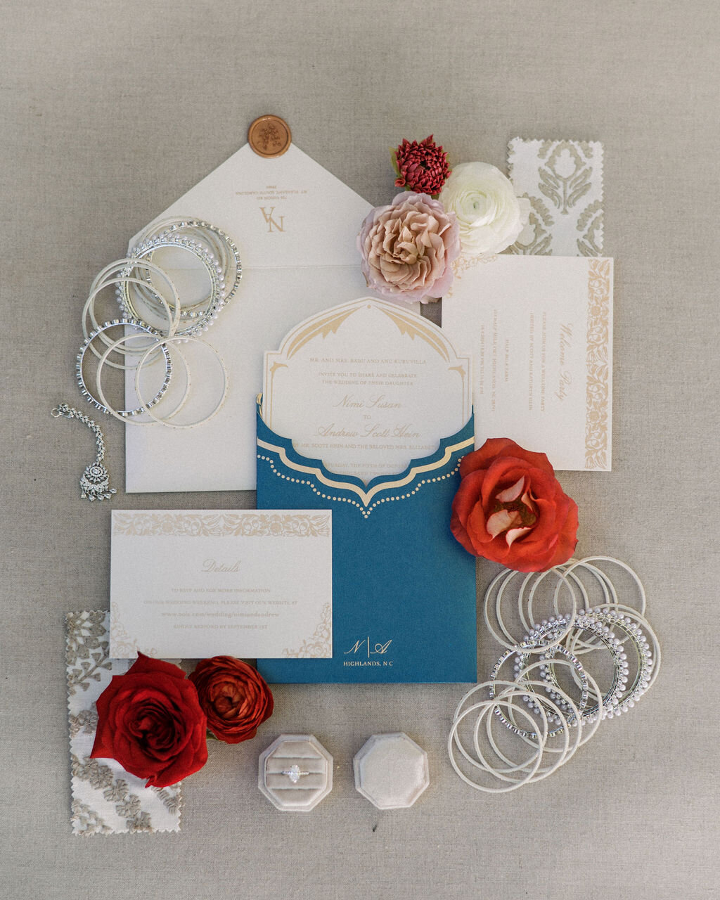 Wedding invitation suite flat lay with blue and gold cards, bangles, floral accents, and ring boxes.