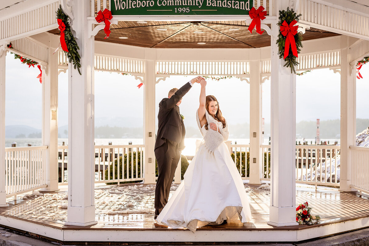 Groom twirling bride in gazebo with winter lake sun shining on them