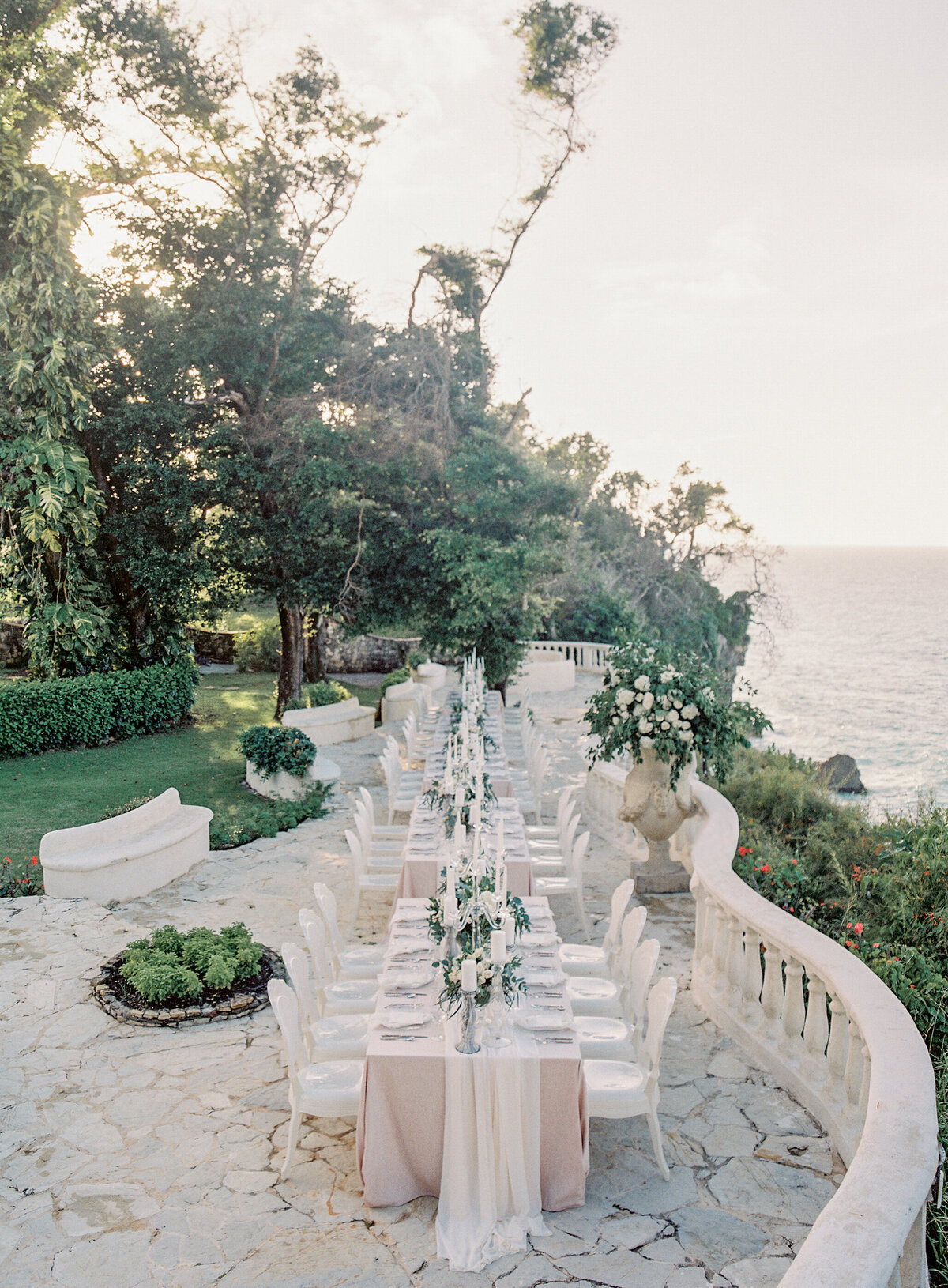 Outdoor wedding reception at top tropical destination wedding venue in the Caribbean, Dominican Republic captured by photographer Laura Gordon. Stone patio overlooking the ocean with long tables decorated with white linens, greenery centerpieces, and elegant table settings. White chairs, lush greenery, and picturesque view complete the scene.